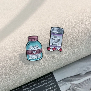 Whimsical enamel pill bottle pins featuring Take Care and Crazy Pills text on metal badges