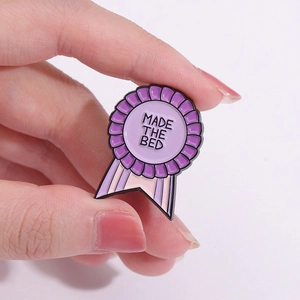 Gros plan d'une broche en émail en forme de médaille avec texte 'Made The Bed' et rubans