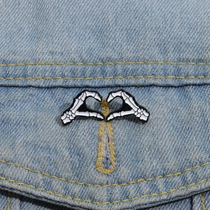 Detail von Skeletthänden als Herzform-Emaille-Pins auf einer hellblauen Jeansjacke