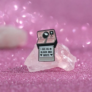 Macro detail of pink retro camera enamel pin with I SEE US IN BLACK AND WHITE text