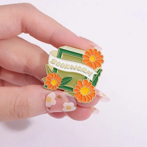 Cartoon enamel pin featuring green bookworm book and orange daisy flowers design