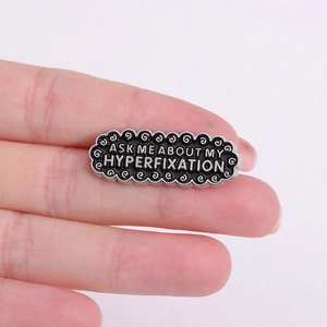 Wavy black enamel brooch with silver Ask Me About My Hyperfixation text held by hand