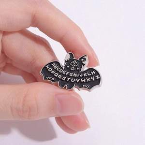 Punk bat enamel pin featuring Ouija board alphabet and wings on a pink background
