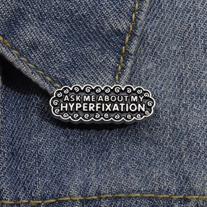 Close-up of black enamel badge with ASK ME ABOUT MY HYPERFIXATION text on denim
