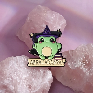 Enamel wizard frog pin with magic wand and spell scroll on a soft pastel background