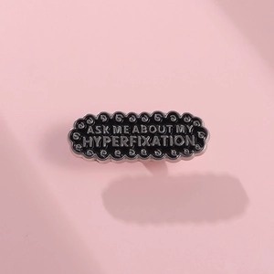 Macro shot of wavy black enamel brooch with white Ask Me About My Hyperfixation text