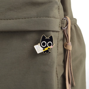Close-up of black cat enamel pin holding a pencil with Keep Trying text on fabric