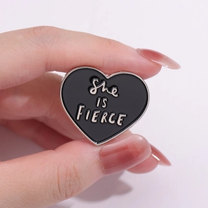 Minimalist black heart enamel pin featuring silver She IS FIERCE text held by hand