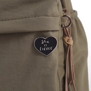 Close-up of black heart enamel pin with She is FIERCE text on a green canvas bag