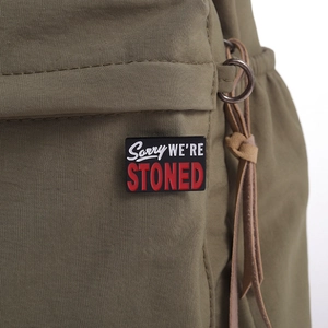Detail der Sorry WE'RE STONED Emaille-Anstecknadel auf einer olivgrünen Segeltuchtasche
