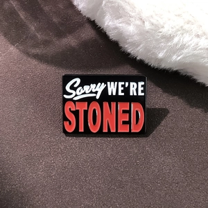 Detail einer rechteckigen schwarzen Emaille-Anstecknadel mit dem Text Sorry We Are Stoned