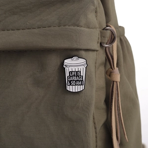 Detail of trash can enamel pin with 'LIFE IS GARBAGE' slogan on a green canvas bag