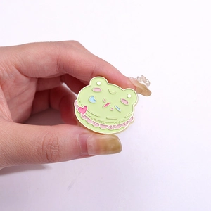 Lindo pin de esmalte de rana macarrón verde con corazones rosas sostenido en la mano