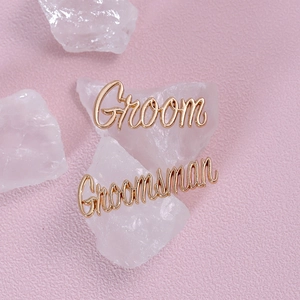 Close-up of rose gold cursive Groom and Groomsman pins on white crystal stones