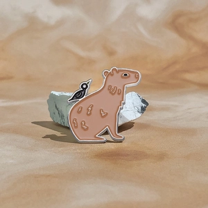 Detailed enamel pin showing a brown capybara with a small black bird on its back