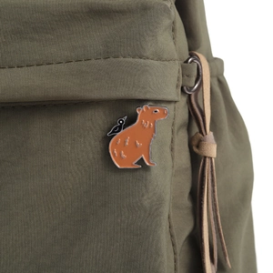 Detail of capybara and bird enamel pin attached to a green canvas backpack
