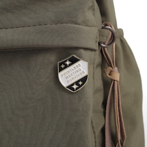 Macro detail of 'Pointless Meeting Survivor' enamel shield pin on green fabric bag