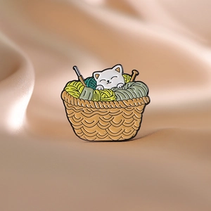 Makroaufnahme einer Emaille-Brosche mit Katze und Wollkorb auf seidigem Stoff