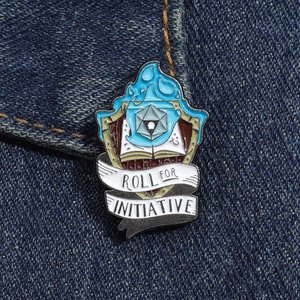 Broche en émail avec dé D20 et grimoire posée sur un tissu en denim bleu foncé robuste