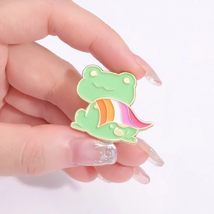 Cute frog enamel pin badge with colorful stripes cape held by hand on light background