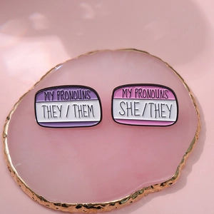 Minimalist enamel pronoun badges with She/They and They/Them text on pink agate tray