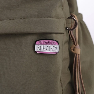 Macro shot of SHE/THEY pronoun enamel pin on a green fabric texture detail