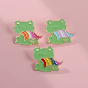 Detailed view of three cute frog enamel pins with colorful striped capes on pink background
