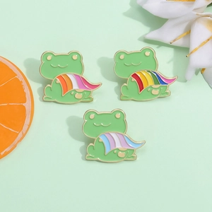 Three cute green enamel frog pins with colorful rainbow capes on a light green background