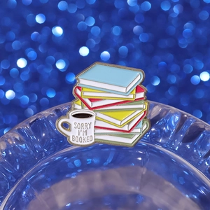 Enamel book and coffee cup pin with 'SORRY I'M BOOKED' slogan on a dark bokeh background