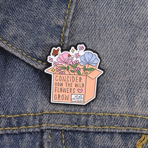 Detail of flower box enamel pin with Luke 12:27 quote on navy blue denim fabric