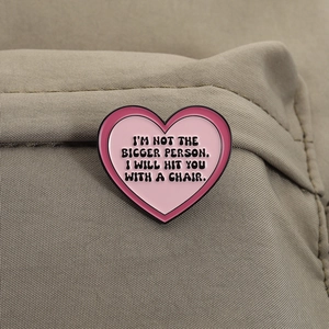 Close-up of pink heart enamel pin with witty slogan on khaki textured fabric