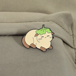 Detail of a cartoon cat enamel pin with a lotus leaf hat on olive green fabric