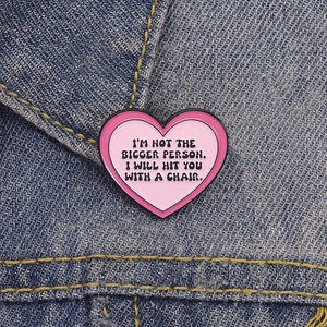 Detail of pink heart enamel pin with quote on denim, showcasing high-gloss finish