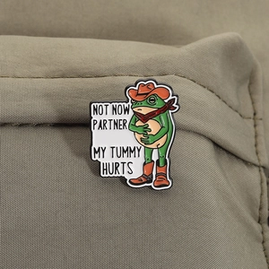 Close-up of funny cowboy frog enamel pin with tummy hurts text on canvas fabric