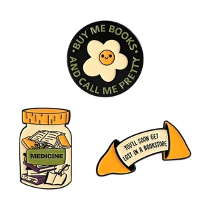 Three book-themed enamel pins including 'Buy me books' and floral designs on white background