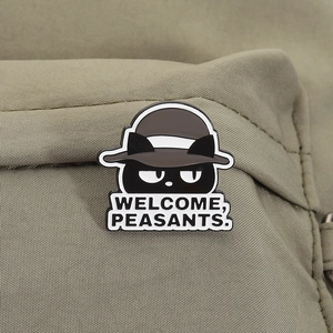 Close-up of a funny black cat enamel pin with Welcome Peasants text on a canvas bag