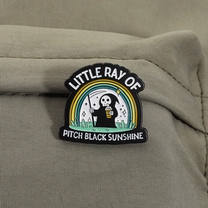 Close-up of enamel reaper pin with Little Ray of Pitch Black Sunshine text on green fabric
