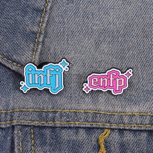 Blue INFJ and pink ENFP enamel pins with gothic text on dark denim fabric background