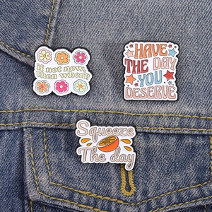 Three enamel pins with inspirational quotes and orange design on denim jacket fabric
