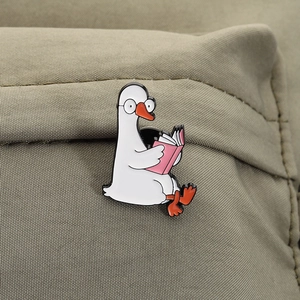 Close-up of a cartoon goose reading a book enamel pin on a green canvas fabric