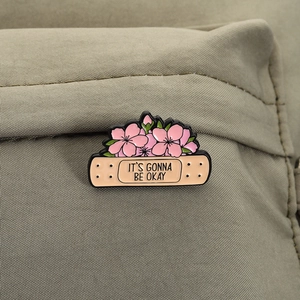 Detail of band-aid enamel pin with pink cherry blossoms and It's Gonna Be Okay text