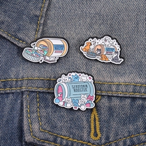 Three creative serotonin booster enamel pins featuring cats and books on denim jacket