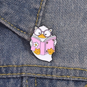 Detail of cute white duck enamel pin with pink book pinned on blue denim fabric