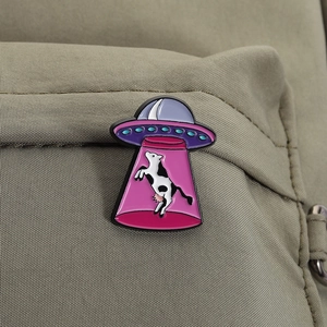 Detailed close-up of a pink UFO and cow enamel pin on olive green fabric texture