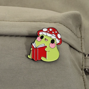 Detailed view of frog enamel pin wearing mushroom hat reading book on canvas bag