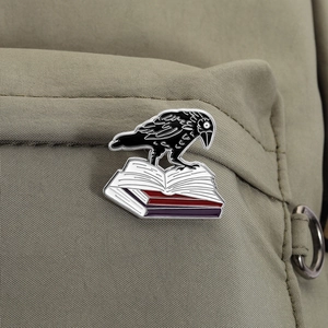 Détail du pin's émaillé corbeau sur livres fixé à un sac à dos en toile kaki