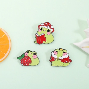 Three creative crown frog enamel pins set against a fresh green background with decor