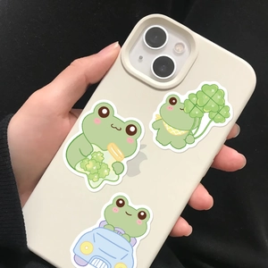 Three cute green cartoon frog stickers applied to the back of a white smartphone