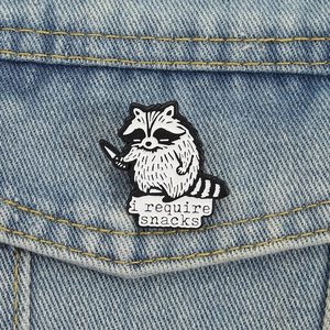 Funny raccoon enamel pin holding a knife with 'i require snacks' text on denim background