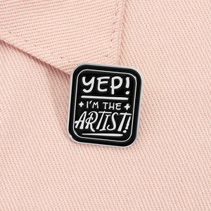 Macro shot of rectangular enamel pin with 'YEP! I'M THE ARTIST!' text on pink fabric